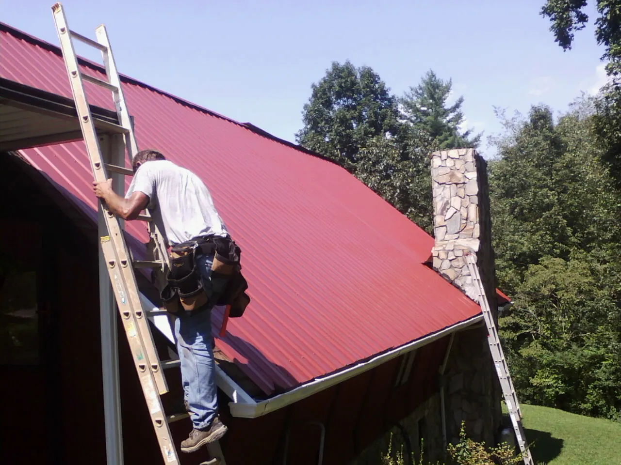 Roofer on a ladder beside a red metal roof and stone chimney — Soffit & Fascia Repair by Roofing Brothers in Grand Saline